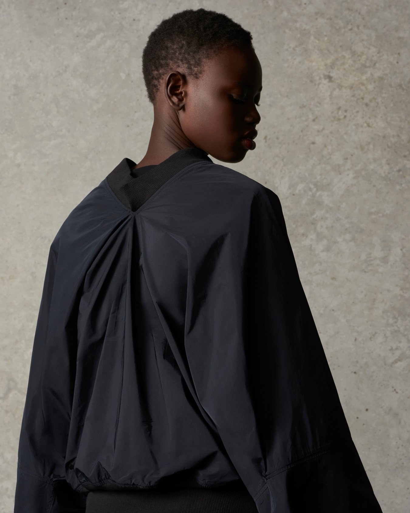 A model seen from behind in a black jacket with a gathered back and ribbed collar, head turned to the side, against a concrete wall