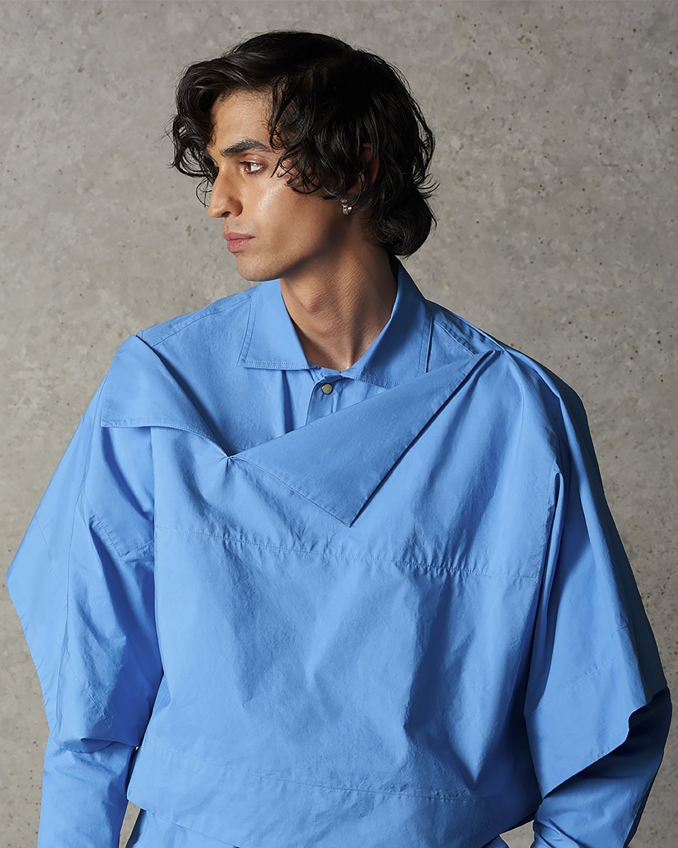A model in a bright blue shirt with an angular layered cut, against a concrete wall